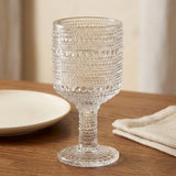 A clear textured glass goblet with a footed stem, featuring horizontal rows of raised circular dots, shown on a wooden table with a plate and napkin in a softly lit indoor setting.