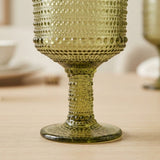A close-up of a textured olive green glass goblet with a dotted pattern, resting on a light wooden table.