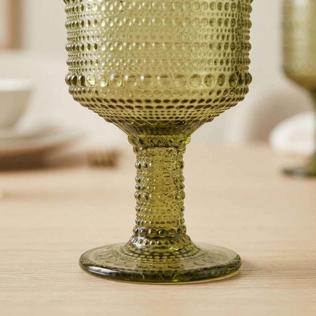 A close-up of a textured olive green glass goblet with a dotted pattern, resting on a light wooden table.