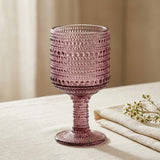 A pink textured glass goblet with a flared base, standing on a beige tablecloth next to a folded napkin and small white flowers.