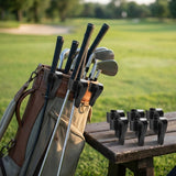 A golf bag on a grassy golf course with several golf clubs inserted, plus a set of black club head rests placed on a wooden table beside it.