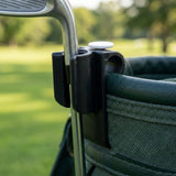 Close-up of a black golf bag clip holding a metal golf club shaft next to a dark green golf bag, with a blurred outdoor golf course background.