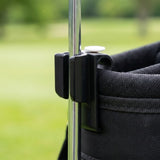 Close-up of a black plastic accessory attached to a silver metal pole and a black fabric bag, with a blurred green outdoor background.