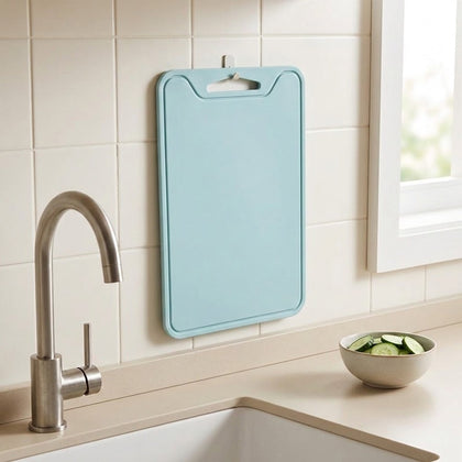 A light blue plastic cutting board hangs on a tiled kitchen wall above a sink with a faucet and a small bowl of sliced cucumbers.