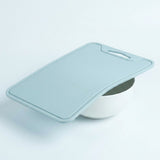 A light blue silicone cutting board with a built-in handle, shown resting on top of a white bowl against a clean white background.