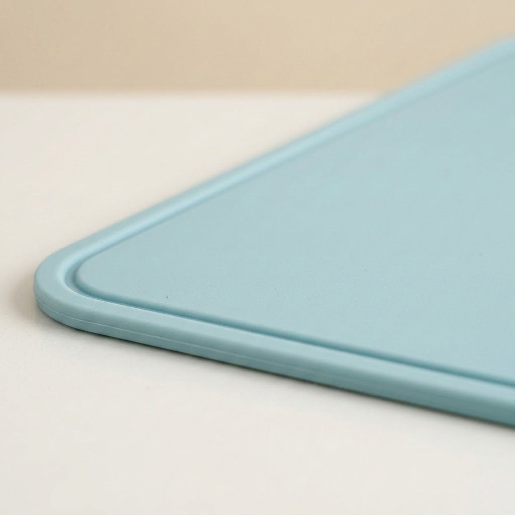 A close-up of a light blue cutting board showing its rounded corner and smooth matte surface, resting on a white surface with a beige background.