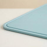 A close-up of a light blue cutting board showing its rounded corner and smooth matte surface, resting on a white surface with a beige background.