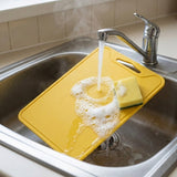 A yellow silicone cutting board rests in a stainless steel kitchen sink under running water, with a sponge on top.
