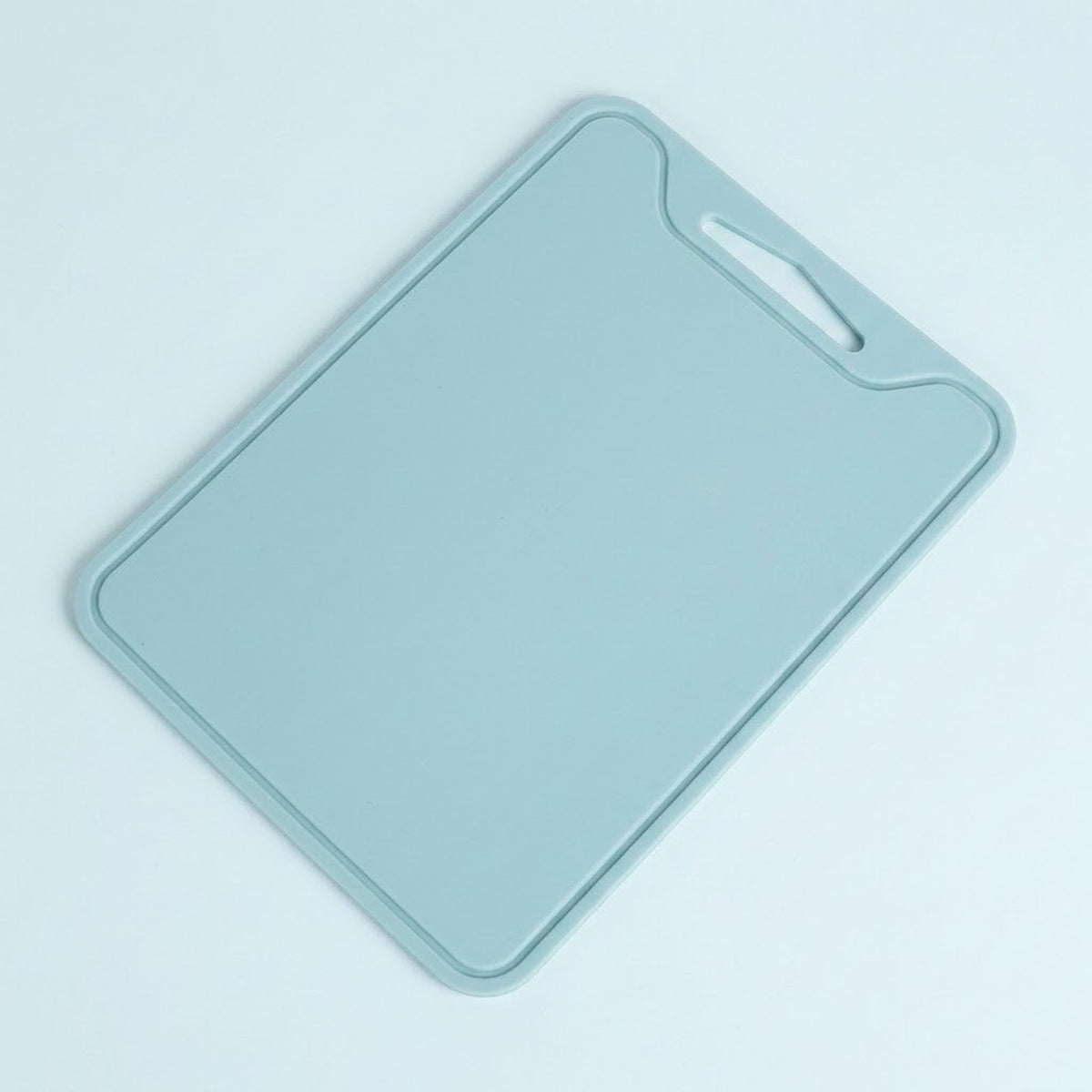 A light blue-gray silicone cutting board with a built-in handle cutout in the top right corner, shown on a clean, light blue background.