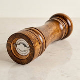 A wooden pepper mill grinder with a polished medium-brown wood body and a silver metal dial, resting on a light beige marble surface.