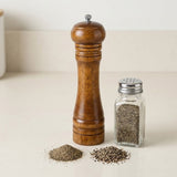 A wooden pepper mill with a silver top knob stands next to a clear glass shaker bottle filled with coarse black pepper, with small piles of ground pepper in front on a light beige countertop.