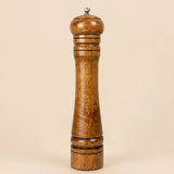 A tall wooden pepper mill with a polished medium-brown wood finish and a small silver metal top knob, set against a solid beige background.