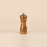 A classic wooden pepper mill grinder with a polished brown wood body and a small silver metal knob on top, set against a solid beige background.