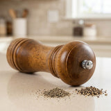 A wooden pepper mill grinder with a silver metal adjustment knob, lying on a light beige countertop with coarse salt and pepper scattered in front, against a blurred kitchen background.