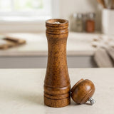 A wooden salt or pepper mill with a matching wooden top placed beside it on a light countertop, with a softly blurred kitchen background.