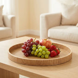 A round light wood serving tray on a matching wooden table, styled with red and green grapes, a red apple, and strawberries in a bright, modern living room setting.