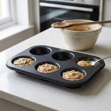 A black six-cup muffin tray filled with batter sits on a light countertop in a kitchen setting, with a mixing bowl and wooden spoon in the background.