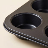 Close-up of a dark non-stick six-cup muffin or cupcake pan on a light beige background.