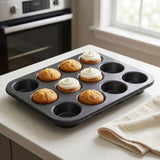 A 12-cup dark gray non-stick muffin tin holding several baked cupcakes, set on a white kitchen countertop near a window and stove.