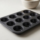 A black 12-cup muffin tin tray on a light gray surface, with a white ceramic bowl partially visible in the background.