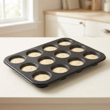 A 12-cup black muffin tin filled with pale batter, placed on a light countertop in a softly blurred kitchen setting.