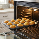 A dark gray non-stick muffin tin with 12 golden-brown baked muffins, shown being pulled out of an oven on a metal rack.
