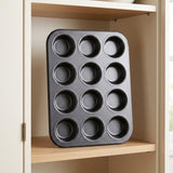 A dark gray non-stick 16-cup muffin tin resting on a light wood shelf inside a beige kitchen cabinet.