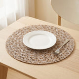 A round woven placemat in beige and white sits on a light wood table, styled with a white plate and a silver fork.