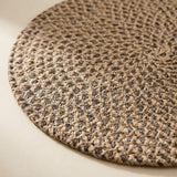 A close-up of a round woven placemat or rug in a braided jute pattern, showing a mix of natural tan and dark brown fibers on a light beige surface.