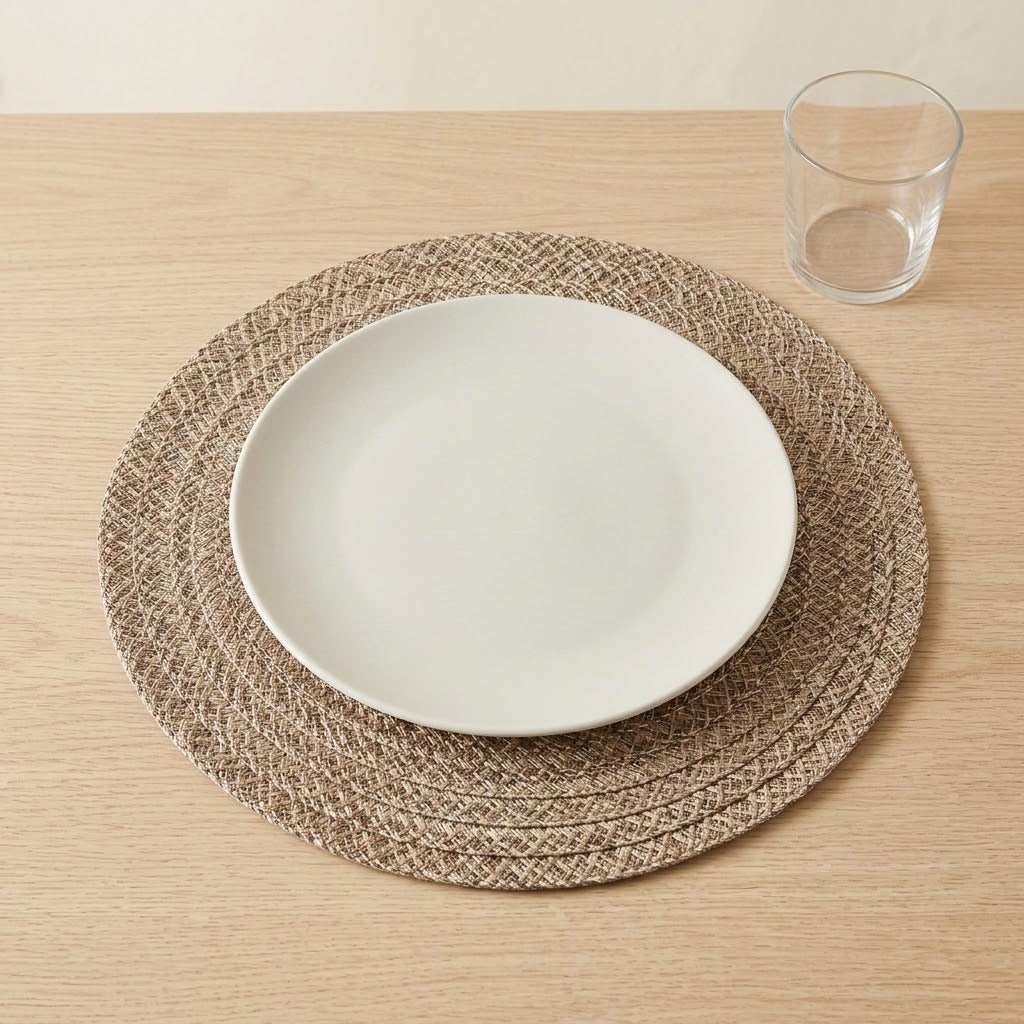 A round woven beige placemat with a white oval plate on top, set on a light wood tabletop next to a clear drinking glass.