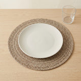 A round woven beige placemat with a white oval plate on top, set on a light wood tabletop next to a clear drinking glass.