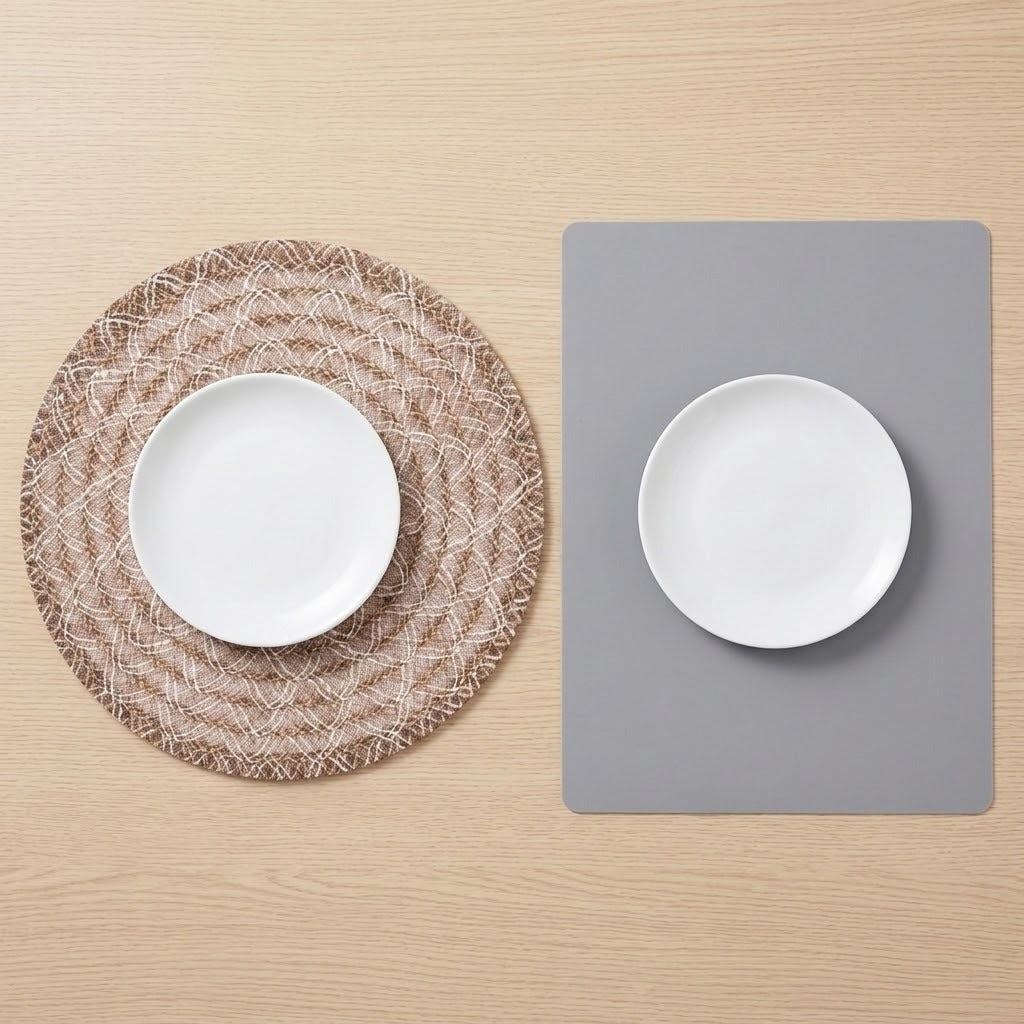 Two place mats on a light wood tabletop, each holding a white plate. One placemat is round with a woven tan-and-white pattern, and the other is a simple gray rectangular mat.
