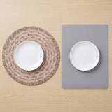 Two place mats on a light wood tabletop, each holding a white plate. One placemat is round with a woven tan-and-white pattern, and the other is a simple gray rectangular mat.