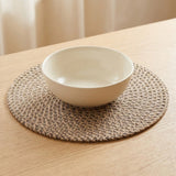 A white ceramic bowl sits on a round woven placemat on a light wood table, with a softly blurred indoor background.
