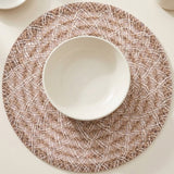 A round woven placemat in beige and white with a textured basketweave pattern, styled with a white ceramic bowl in the center on a light cream tabletop.