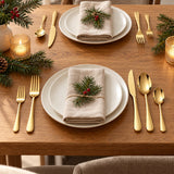 Gold stainless steel cutlery sets a holiday dinner table with plates, linen napkins and evergreen sprigs.