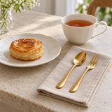 A gold stainless steel teaspoon and tea fork sit beside a teacup and dessert plate on linen.