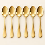 Six gold stainless steel table spoons are aligned on a pale studio backdrop.
