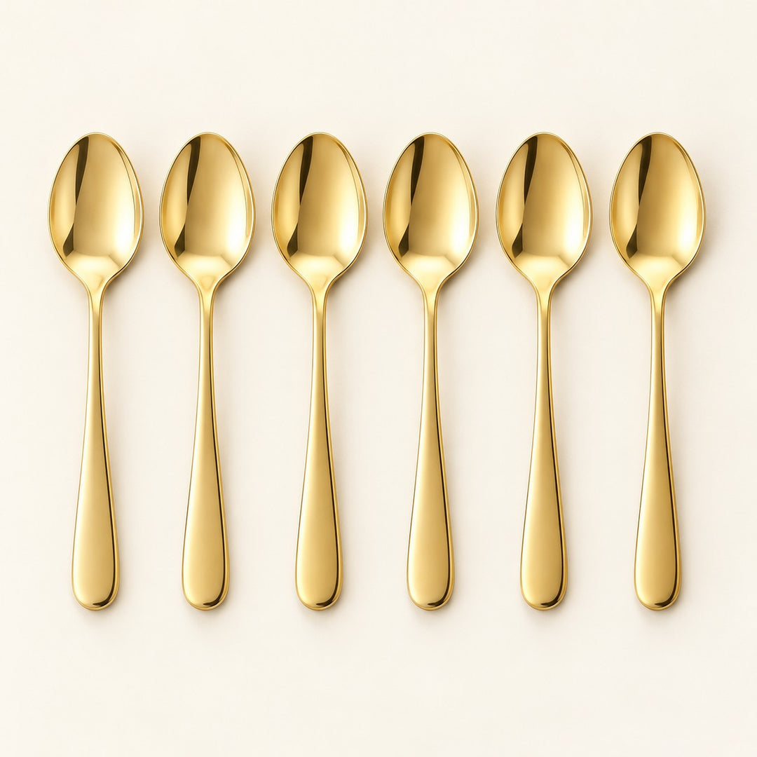 Six gold stainless steel teaspoons are arranged in a row on a pale studio backdrop.