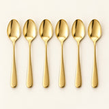 Six gold stainless steel teaspoons are arranged in a row on a pale studio backdrop.