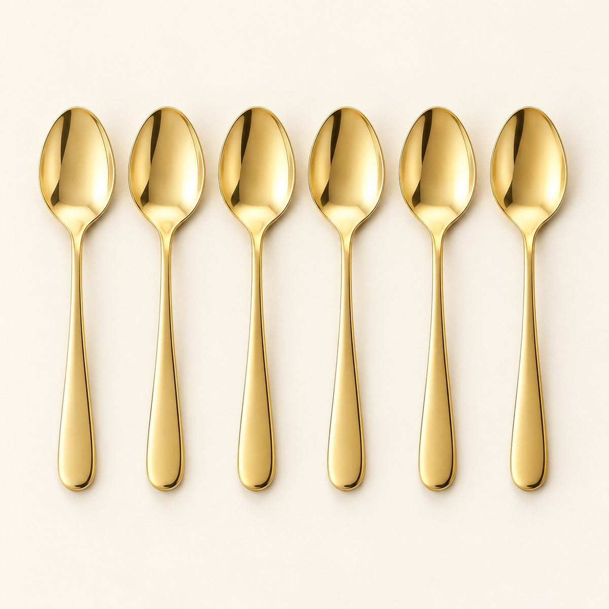 Six gold stainless steel teaspoons are arranged in a row on a pale studio backdrop.