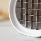 Close-up of a white kitchen utensil with a metal grid cutting or straining element, photographed on a light neutral surface with a blurred background.