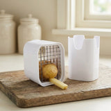 A white plastic french fry cutter and holder set on a rustic wooden cutting board, with a potato and a fry visible inside the cutter. The background shows a softly blurred kitchen setting with cream-colored jars and a window.