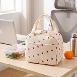 A cream-colored insulated lunch bag with a strawberry print sits on a light wood desk in a bright office setting, next to a laptop, a container of vegetables, an orange, and a stainless steel bottle.