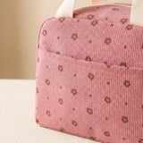 A close-up of a pink textured fabric pouch or lunch bag with a repeating brown floral dot pattern and light beige handles, resting on a light surface against a soft beige background.