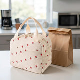 A cream-colored insulated lunch bag with a red strawberry pattern sits on a kitchen counter next to a brown paper bag. The background shows a softly blurred kitchen interior with a coffee maker and microwave.