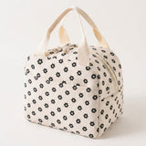 An off-white canvas lunch bag with a black floral pattern and cream-colored handles, photographed on a clean white background.