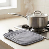 A gray quilted pot holder with a loop handle rests on a beige kitchen countertop next to a stainless steel pot on a gas stove with steam rising.