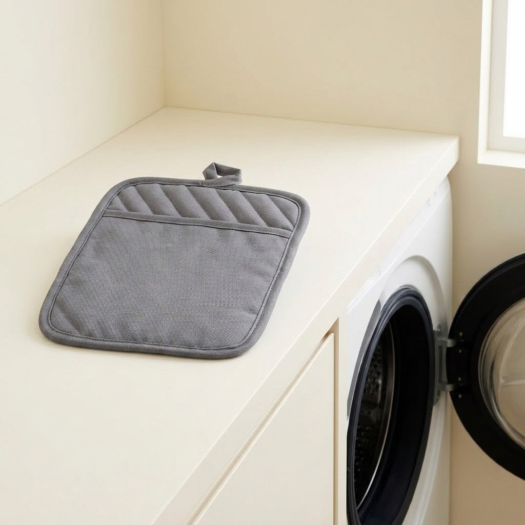 A gray quilted cotton pot holder with a loop handle rests on a cream-colored laundry counter next to a white washing machine with its door open.