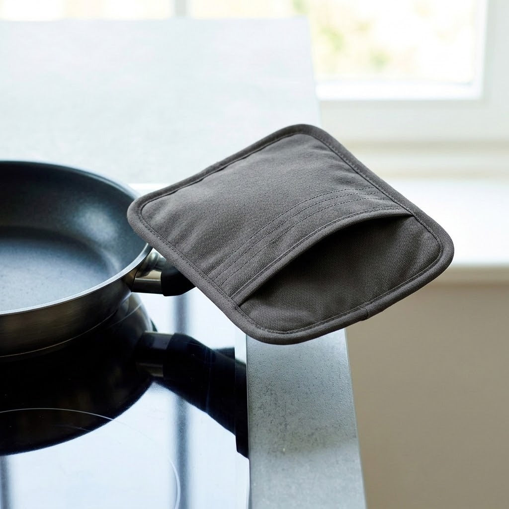 A dark gray fabric kitchen pot holder with a front pocket, draped over the edge of a stovetop next to a black frying pan.